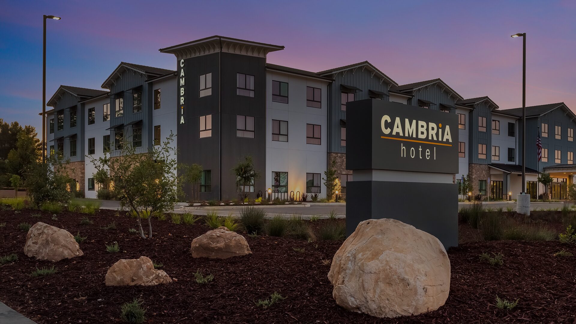 Exterior view of Cambria Hotel at dusk with illuminated sign and landscaping.