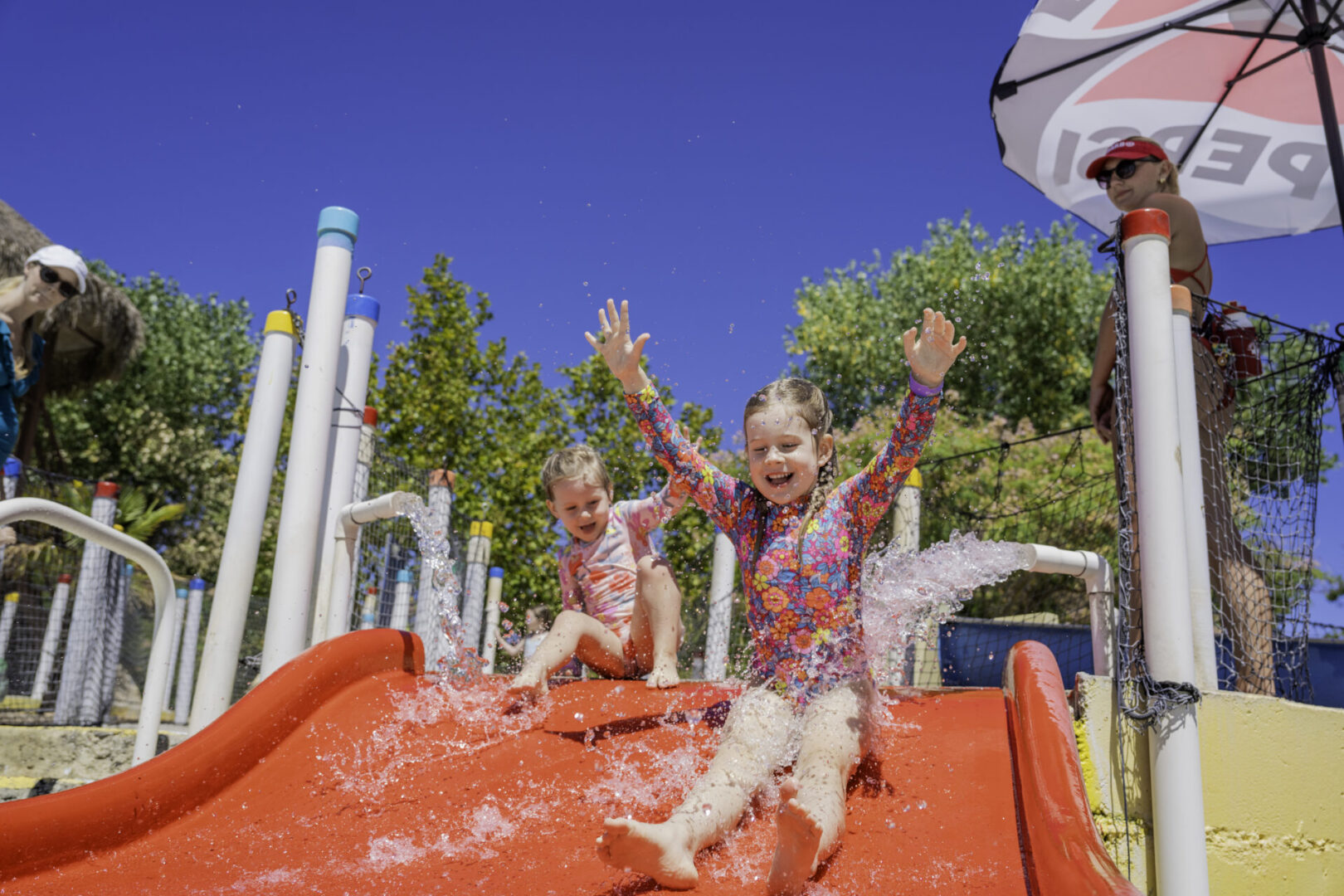 Two children joyfully sliding down a water slide on a sunny day.